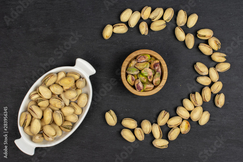 Lot of whole ripe green pistachio in tiny wooden bowl in white oval ceramic bowl flatlay on grey stone