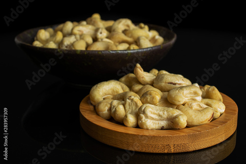 Lot of whole unsalted beige cashew on round bamboo coaster in dark ceramic bowl isolated on black glass