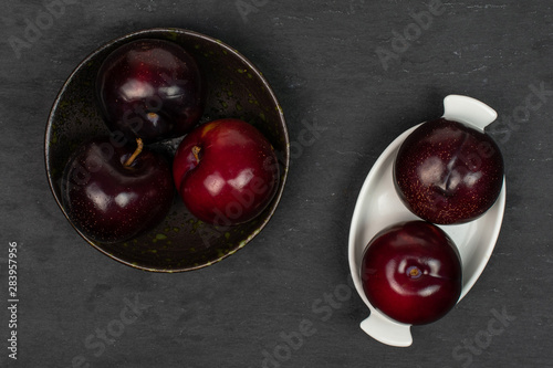Group of five whole ripe red round plum in dark ceramic bowl in white oval ceramic bowl flatlay on grey stone