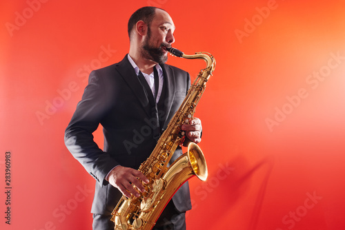 Portrait of professional musician saxophonist man in  suit plays jazz music on saxophone, red background in a photo studio