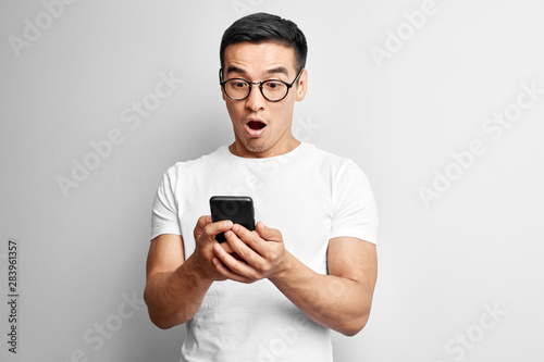 Handsome Asian guy in white studio looks surprised at screen of mobile phone. Shocked Kazakh man in glasses dressed casual holds smartphone in hands