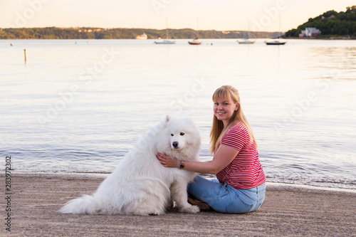 Horizontal side view of pretty smiling blond young woman sitting petting her gorgeous samoyed dog with small sailboats on river in the background