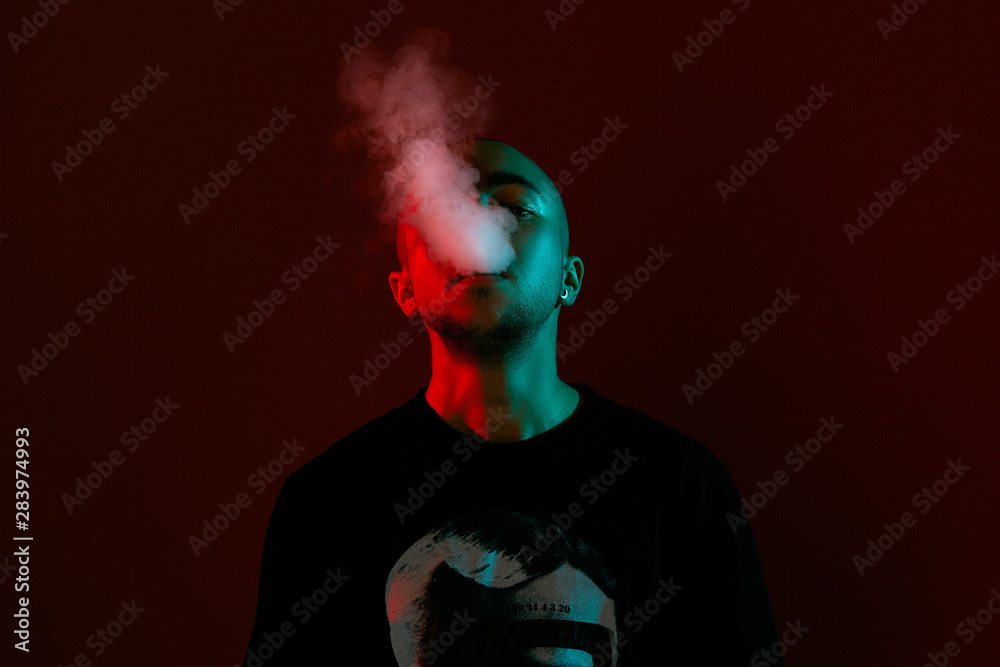 Close-up studio shot of a young bald guy vaping, blowing out a cloud of smoke on red background.