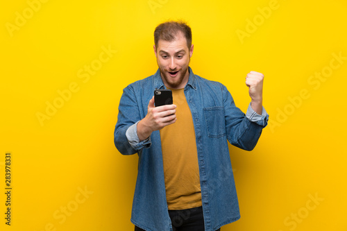 Blonde man over isolated yellow wall surprised and sending a message
