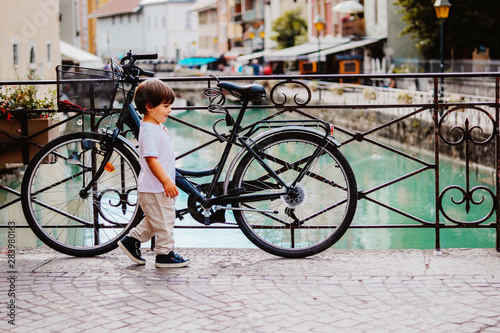 Little boy walking over bridge of Thiou River in Annecy, France near parked bicycle. Child in the city. Little tourist, summer lifestyle. Discovering Europe, traveling with kid.