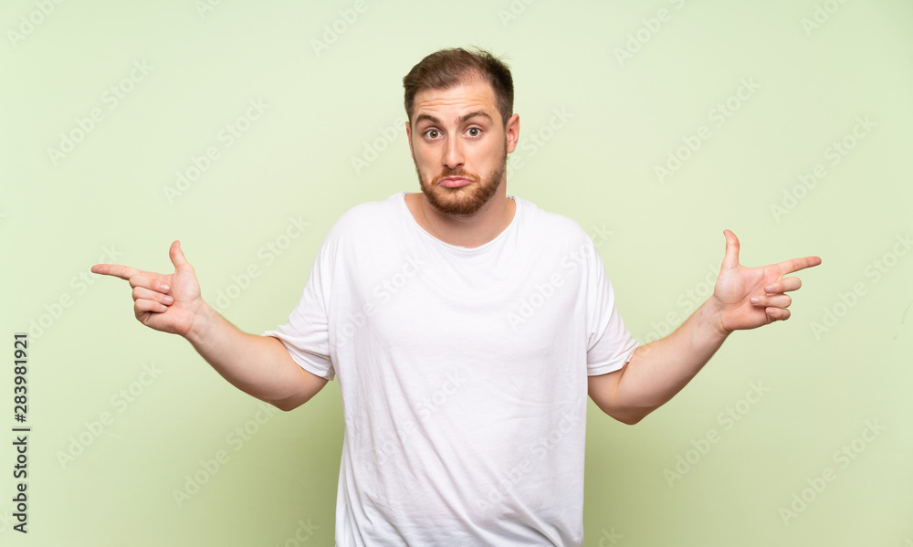 Handsome man over green background pointing to the laterals having doubts Handsome man over green background pointing to the laterals having doubts