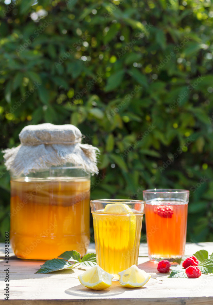 Fermented drink Kombucha in a glass jar and two glasses with a drink with lemon and raspberry in the foreground, in the summer garden, on a wooden table. Fermented drink Kombucha in a glass jar and two glasses with a drink with lemon and raspberry in the foreground, in the summer garden, on a wooden table.