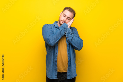 Blonde man over isolated yellow wall making sleep gesture in dorable expression