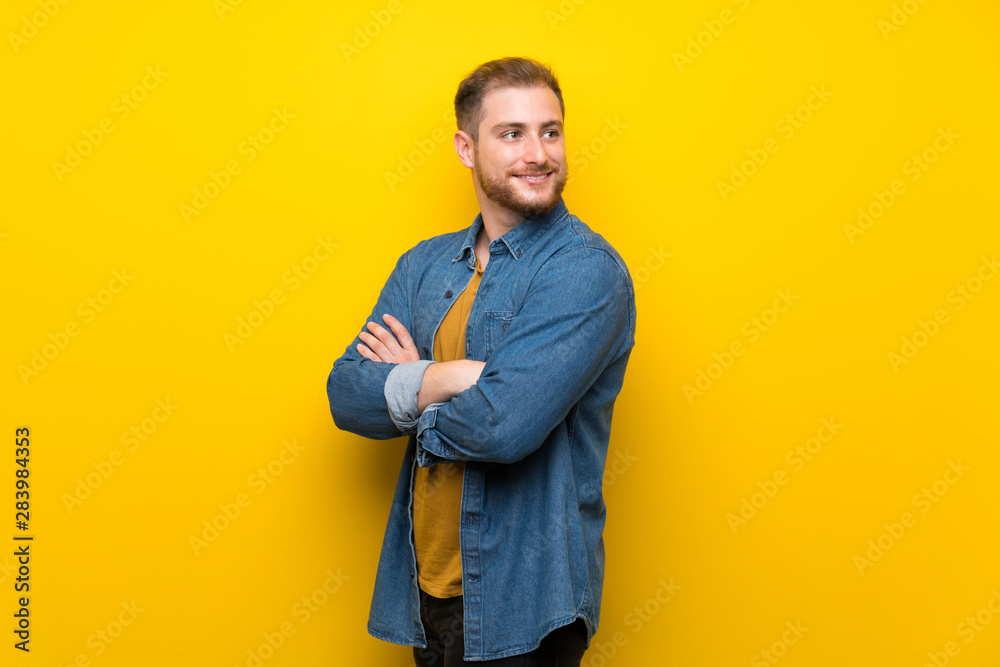 Blonde man over isolated yellow wall with arms crossed and happy