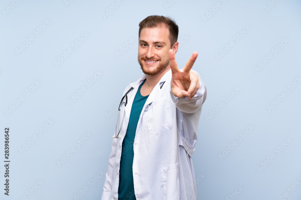 Handsome doctor man over blue wall smiling and showing victory sign Handsome doctor man over blue wall smiling and showing victory sign