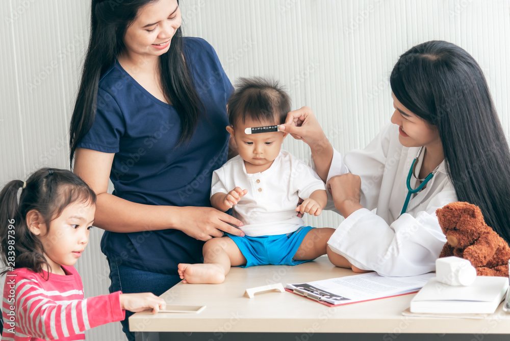 The Asian woman doctor is measuring the temperature of the boy, whose mother took the son and daughter to receive treatment from the flu, Which is pandemic, to health care and health insurance concept The Asian woman doctor is measuring the temperature of the boy, whose mother took the son and daughter to receive treatment from the flu, Which is pandemic, to health care and health insurance concept