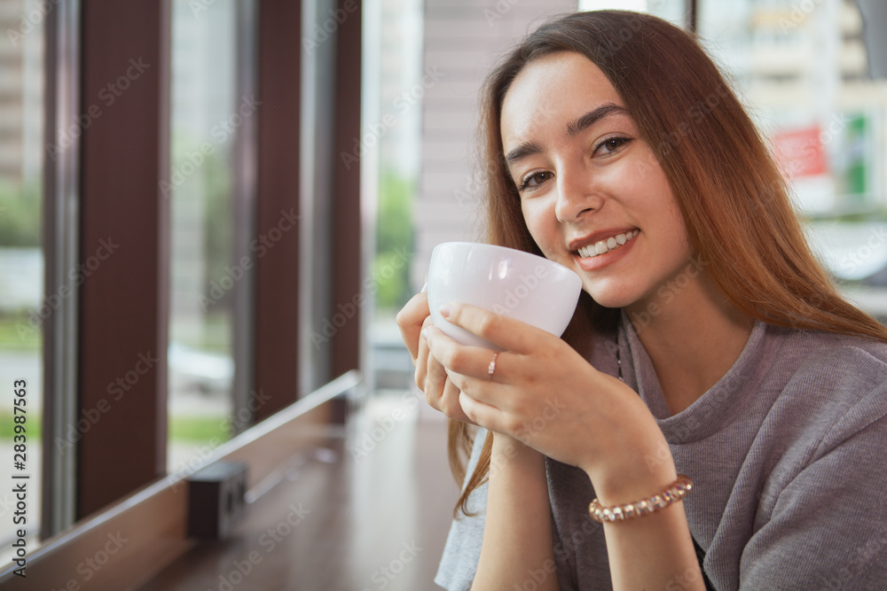 Close up of a lovely young woman smiling to the camera, holding cup of delicious fresh coffee, copy space. Happy woman enjoying morning coffee Close up of a lovely young woman smiling to the camera, holding cup of delicious fresh coffee, copy space. Happy woman enjoying morning coffee