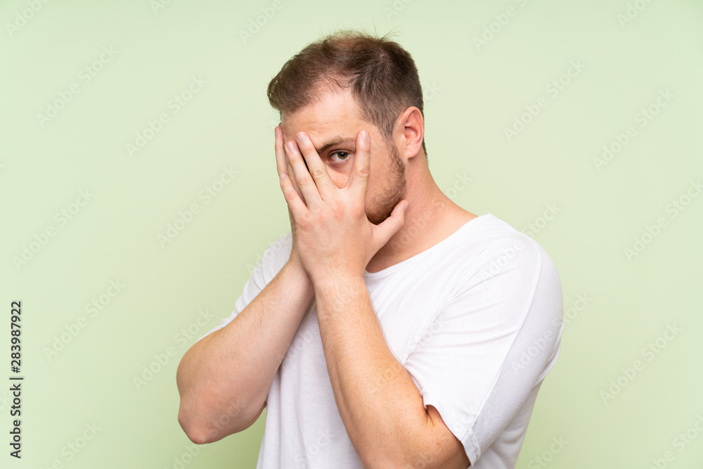 Handsome man over green background covering eyes and looking through fingers Handsome man over green background covering eyes and looking through fingers