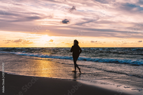 Teenage girl running barefoot on a sandy seaside beach at sunset - a silhouetted photo of a young woman against the sky