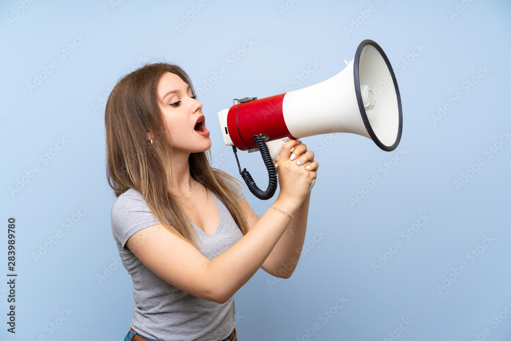 Teenager girl over isolated blue wall shouting through a megaphone Teenager girl over isolated blue wall shouting through a megaphone