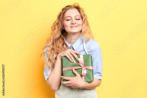 attractive blonde girl enjoying receiving a present, isolated yellow background, stduio shot, happiness, girl hugging a box with gift photo