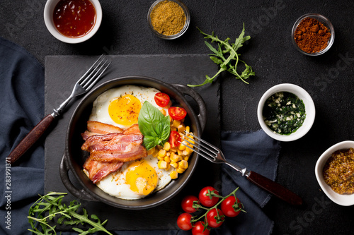 Two fried eggs with bacon, tomatoes and corn in a black pan on a dark background Top view with copy space for your text
