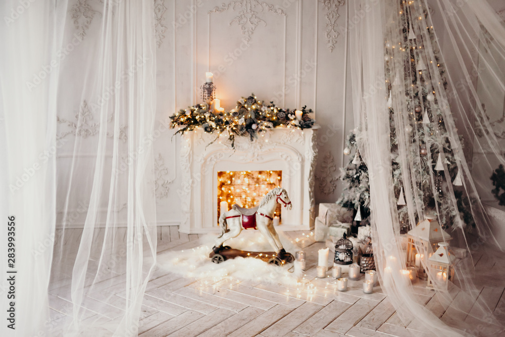 classical luxury interior of a white room with Christmas tree with garland, decorated fireplace, rocking horse, gifts for new year