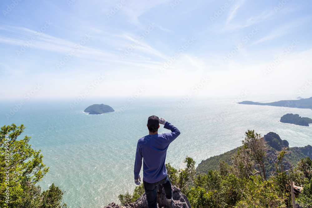 Men in blue shirts are looking at the beauty of the sea on the hilltop with blue sky. Men are looking at the success of climbing.
