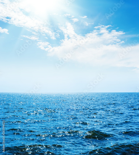 blue sea with waves and sky with sun and clouds over it
