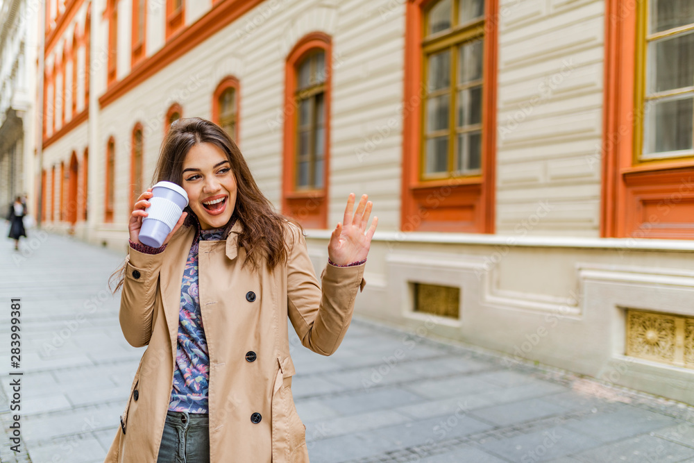 Fototapeta premium Coffee on the go. Beautiful young woman holding coffee cup and smiling while walking along the street. Young beautiful brunette drinking coffee walking around the city.