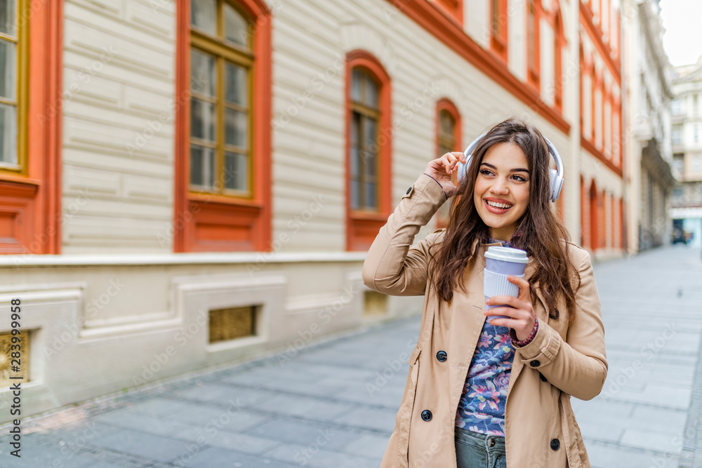 Smiling brunette woman in autumn clothes holding cup of coffee and listening music.