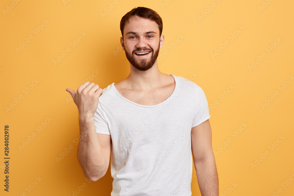 cheerful handsome joyful man pointing with thumb on copyspace behind his back isolated on yellow background. close up portrait. look here, this way, please, direction cheerful handsome joyful man pointing with thumb on copyspace behind his back isolated on yellow background. close up portrait. look here, this way, please, direction