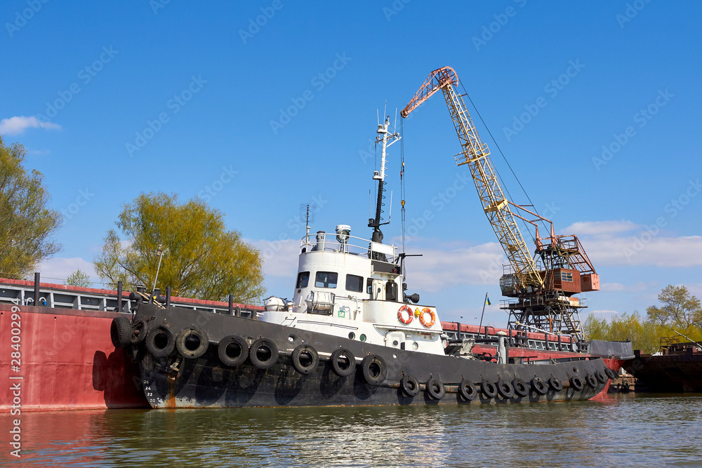 Fototapeta premium Tugboat with row of black tires used as boat bumpers in harbor near port crane