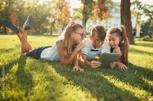 three smiling school-age friends are lying on the grass and looking at the tablet