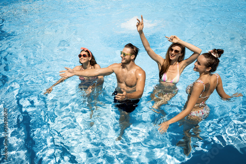 Company of young merry girls and guy are having fun in the swimming pool on the open air on a sunny summer day