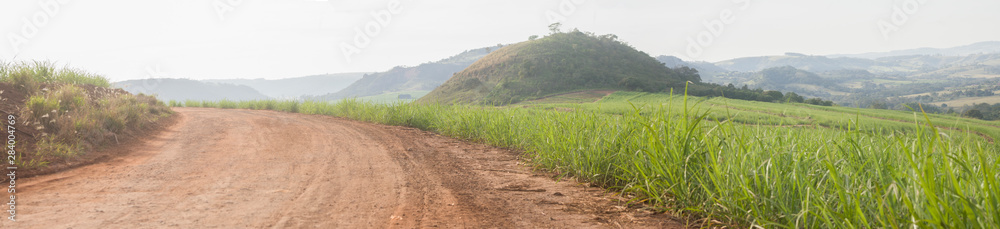 Panoramana de paisagem com estrada rural, mantanhas e plantação de cana de açúcar.