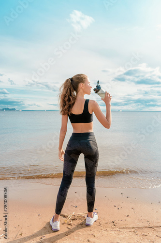 Slender athletic girl in sports clothes drinking water from a bottle on the beach against the beautiful sky