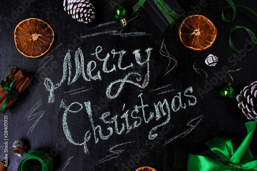Christmas background with a words:Merry christmas, dry orange,white pinecone,green christmas tree balls,ginger sticks,green ribbon on a black background,flat lay