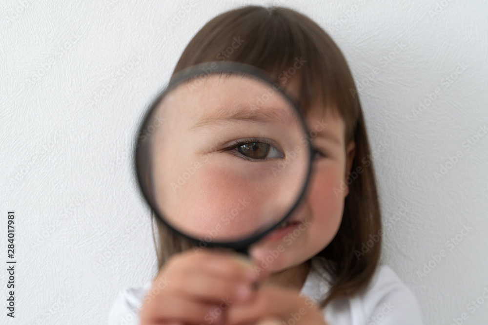 A child is looking through a magnifying glass. The large eye of a child behind a magnifying glass. Little girl at the optometrist. A child is looking through a magnifying glass. The large eye of a child behind a magnifying glass. Little girl at the optometrist.