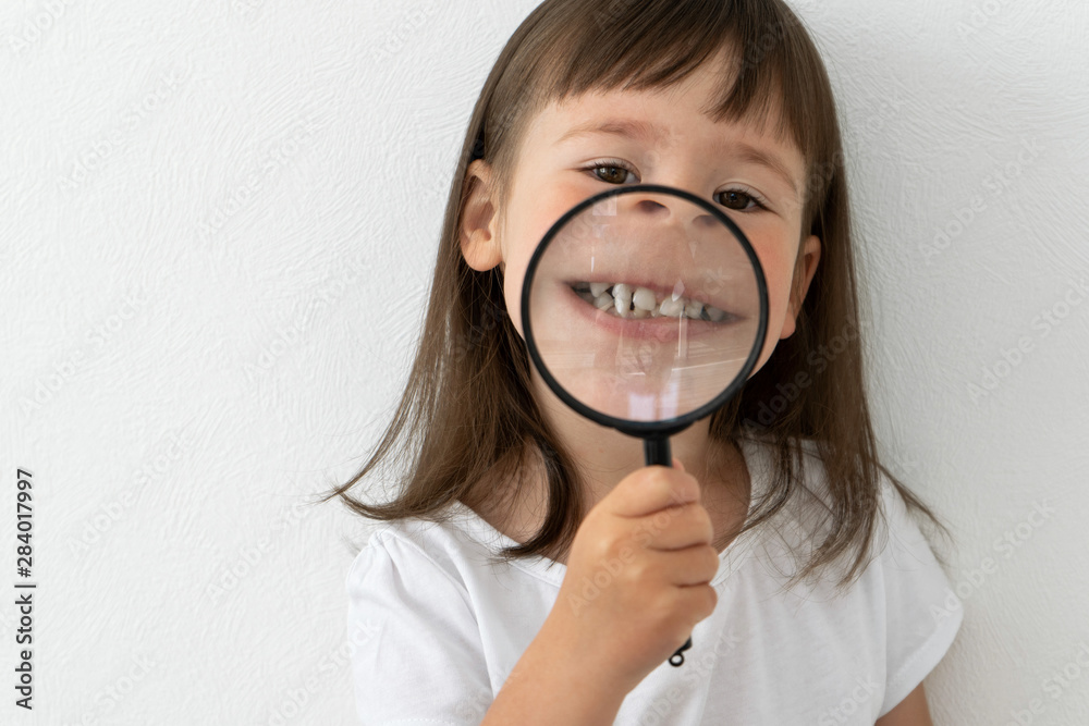 Little girl shows her teeth. Baby teeth through a magnifying glass. The kid holds a magnifier in his hands and shows his teeth. Little girl shows her teeth. Baby teeth through a magnifying glass. The kid holds a magnifier in his hands and shows his teeth.