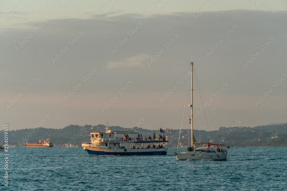 Small pleasure tourist boat sailing from the port for a cruise off the coast on a summer evening against the mountains, Batumi Small pleasure tourist boat sailing from the port for a cruise off the coast on a summer evening against the mountains, Batumi