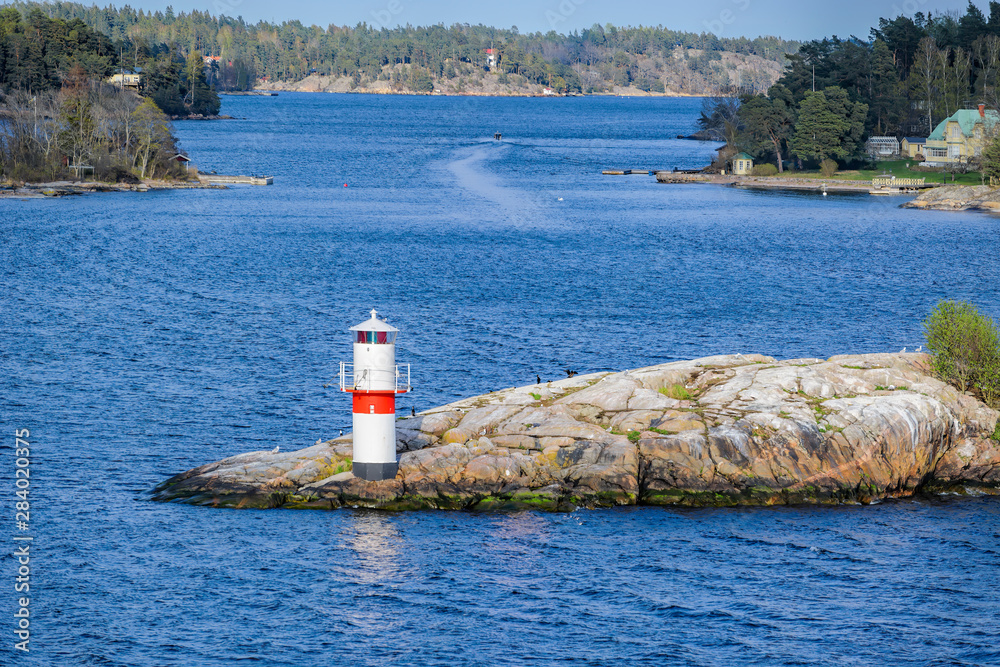 Φωτογραφία Beacon or small lighthouse in red and white at a rocky shore of Oxdjupet strait during the evening with coniferous forest and dwelling islands on the background