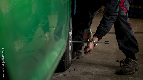 Man changing car tires in garage because of winter season. Car elevated on jack. Workplace environment in dark colors, bright green car.