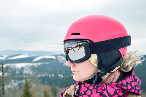 Close up portrait of snowboarder woman at ski resort wearing helmet and goggles with reflection of mountains.