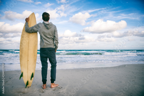 Canvas Print Surfer in gray hoodie holding his surfboard on the shore of a windy tropical bea