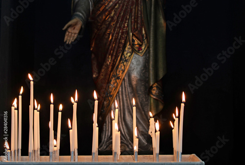 Burning white devotion candles in church from believers to pray and ask for help in front of religious sculpture of Jezus with stigmata on hand and ornate robe
