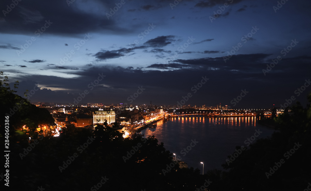 KYIV, UKRAINE - MAY 21, 2019: Beautiful view of night cityscape with illuminated buildings near river and bridge
