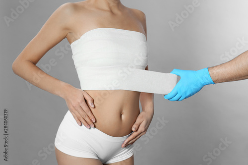 Fotografia Doctor bandaging young woman after plastic surgery operation on grey background,