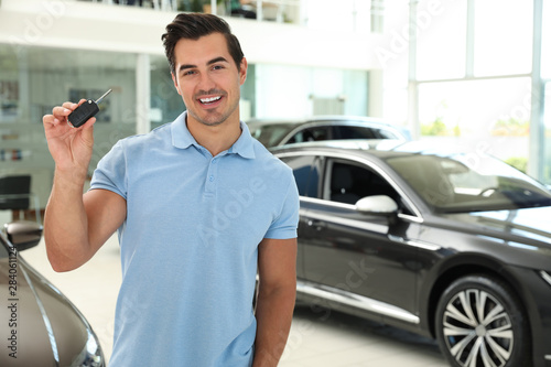Happy young man with car key in modern auto dealership. Space for text