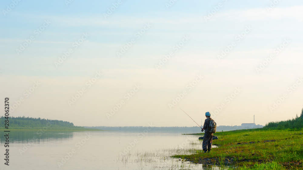 Yakut Asian girl tourist fisherman with a backpack and a cap to fish in the river vilyu in the haze at sunset in the wild North of Russia on the background of the village Suntar.