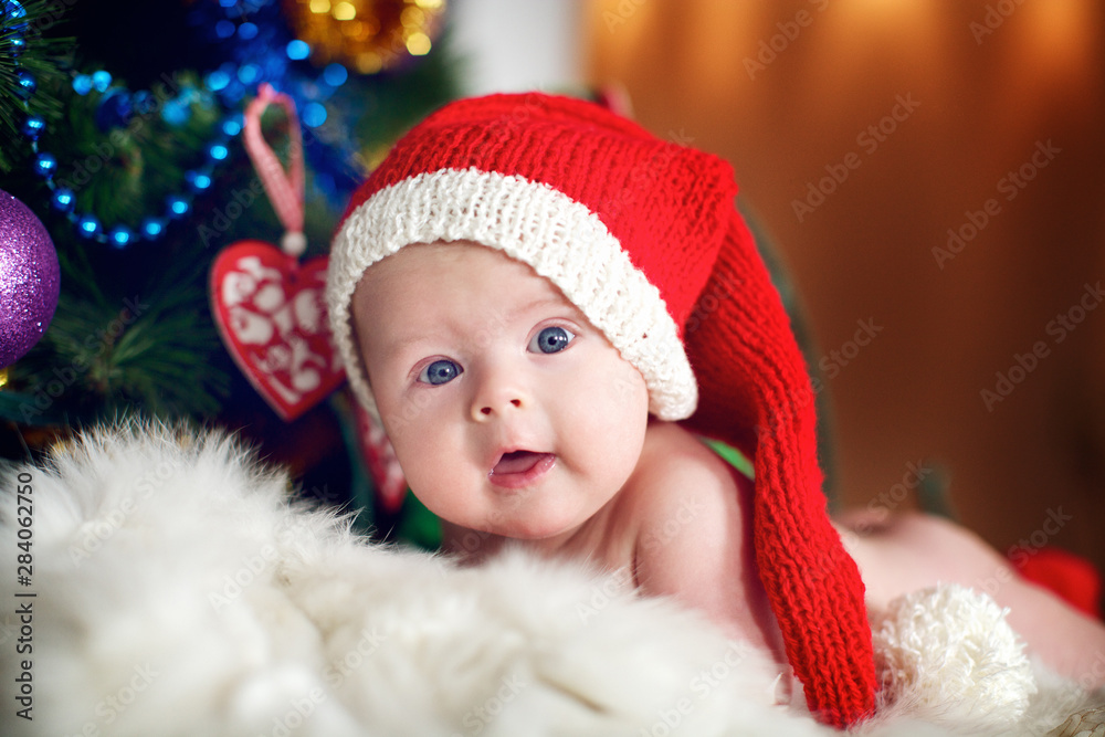 Little baby in red cap of Santa Claus celebrates Christmas. Christmas photo of infant in red cap. New Year's holidays and Christmas tree. Little baby in red cap of Santa Claus celebrates Christmas. Christmas photo of infant in red cap. New Year's holidays and Christmas tree.