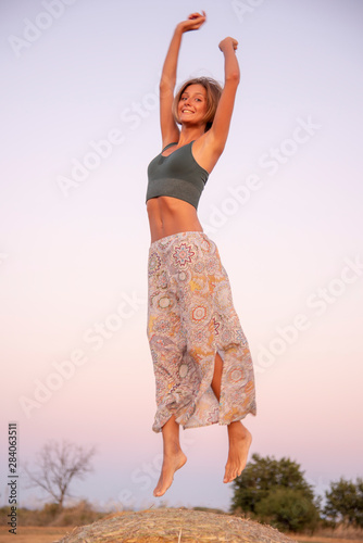 A beautiful  slender  smiling girl raises her hands up and bounces on haystacks in the light of sunset sunlight.