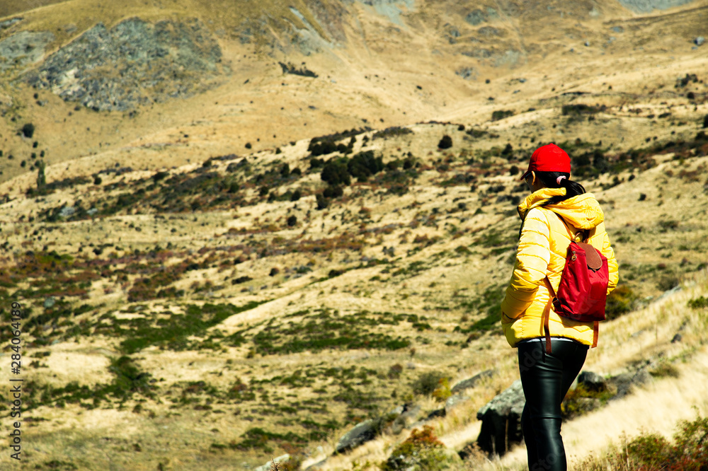  Asian woman tourist with yellow jacket and red backpack look at the beautiful view on the way to Roys Peak  trail in New Zealand.