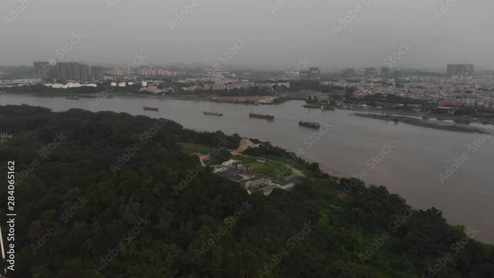 A drone flies over a park area with green trees. Behind the park, a river flows. Body ships sailing on the river A drone flies over a park area with green trees. Behind the park, a river flows. Body ships sailing on the river