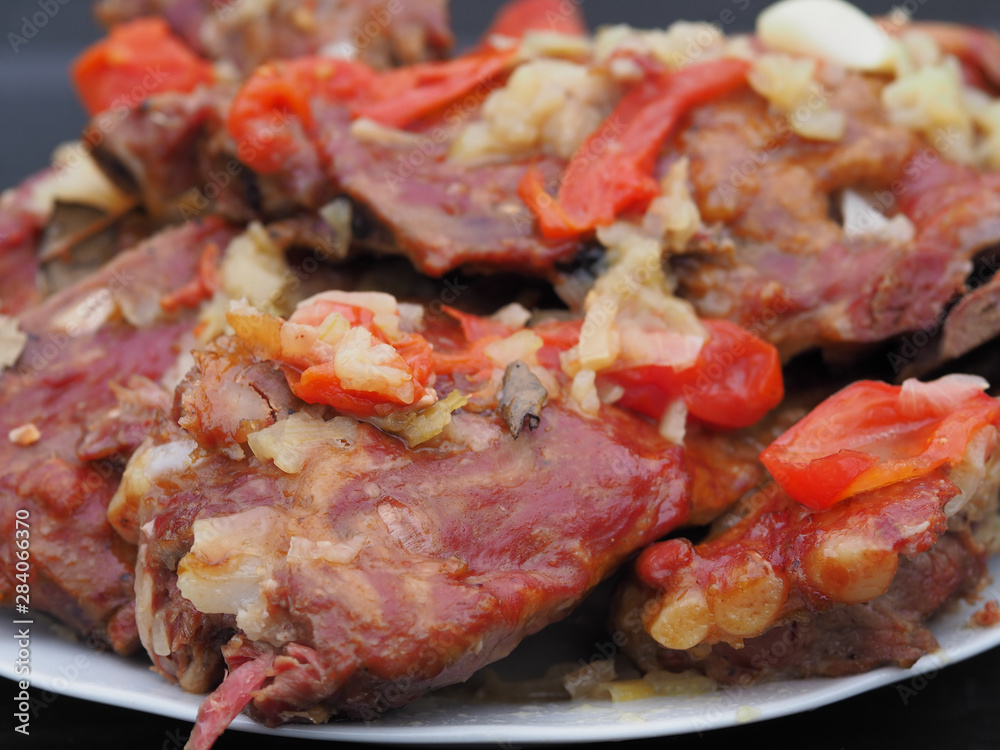 Appetizing and beautiful smoked pork ribs are stewed in a sauce with tomatoes , onions and herbs on a white plate. High-calorie and tasty isolated meat dish close-up